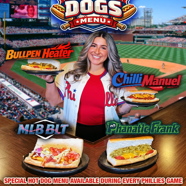 Woman in Phillies jersey holding two hot dogs, with two more on a table, against a baseball stadium backdrop.