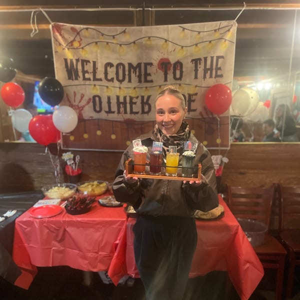 A smiling woman holds a tray of four colorful drinks with whipped cream and themed toppers at Eddington House.