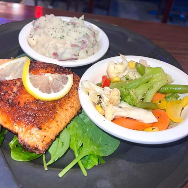 A salmon fillet with lemon, mashed potatoes, and mixed vegetables on a dark plate.