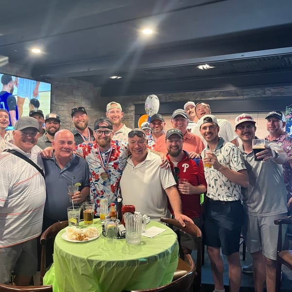 A large group of men smiling and posing for a photo around a table with drinks and snacks.