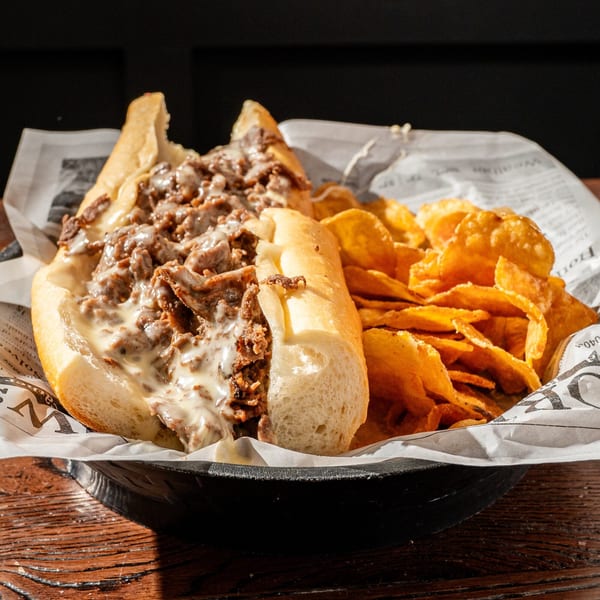 A cheesesteak sandwich with melted cheese and a side of potato chips in a basket.