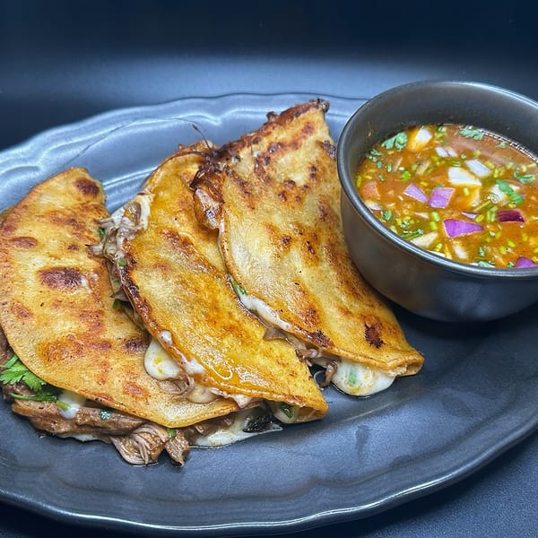 Three cheesy birria tacos with a side of consommé on a dark plate.