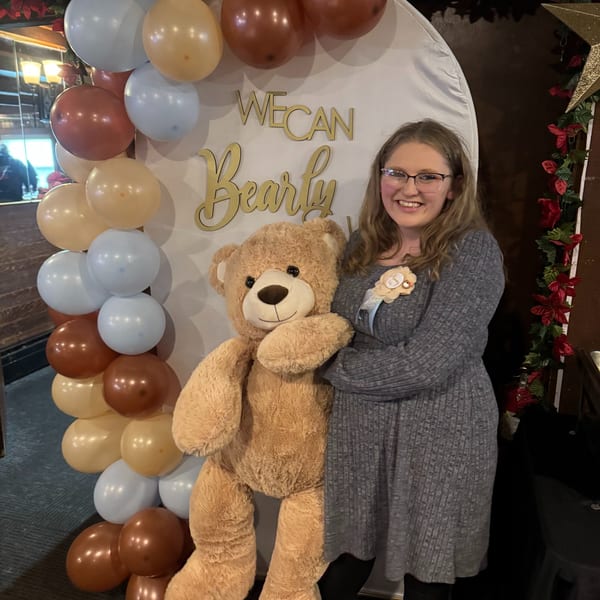 A smiling woman in glasses stands next to a large teddy bear and a balloon arch.