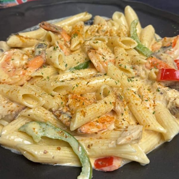 Creamy penne pasta with shrimp, chicken, red and green peppers, and seasoning on a black plate.